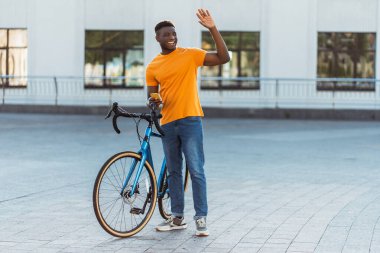 Gülümseyen olumlu Nijeryalı adamın el sallayıp bisikletin yanında cep telefonu tutması. Şehir caddesinde şık sarı tişört giyen yakışıklı bir erkek. Aktif sağlıklı yaşam tarzı kavramı