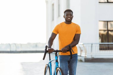 Bisiklet tutan, şehir caddesinde yürüyen pozitif gülümseyen Afrikalı adamın portresi. Yakışıklı, şık günlük kıyafetler giyen, kameraya bakan bir erkek. Sağlıklı yaşam tarzı konsepti, bisiklet