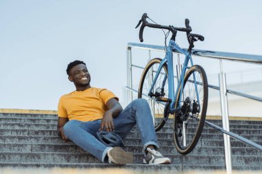 Süslü turuncu giysiler içinde gülümseyen Afrikalı Amerikalı adam merdivenlerde yanında bisikletle oturuyor. Bisiklet sürdükten sonra kaskını takan çekici bir erkek. Sağlıklı yaşam tarzı kavramı