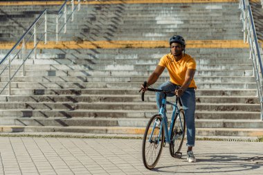 Şehir caddesinde bisiklet süren yakışıklı, gülümseyen, kask takan ve başka yöne bakan Afro-Amerikan adam manzarası. Spor, hobi, aktif yaşam tarzı konsepti 