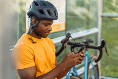 Güvenlik kaskı takan yakışıklı Nijeryalı adamın portresi otobüs durağında oturuyor, cep telefonunu tutuyor, sokakta video izliyor. Akıllı telefon kullanan çekici bir erkek bekliyor. Çevrimiçi alışveriş kavramı