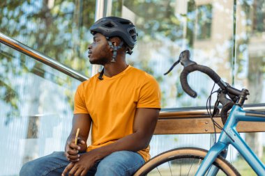 Rahat Afro-Amerikan bir adam, bisiklet sürdükten sonra otobüs durağında dinlenirken, elinde cep telefonu tutarak kafasını çeviriyor. Halk yaşam tarzı konsepti