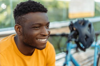 Otobüs durağında oturan yakışıklı Afro-Amerikan adam, yanında bisiklet ve güvenlik kaskı. Sokakta, dışarıda gülümseyen olumlu bir erkek bekliyor. Bisiklet hobisi