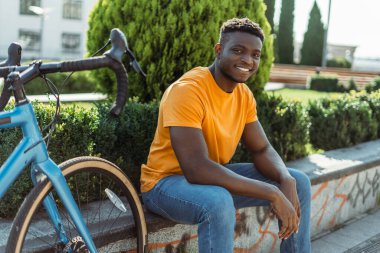 Pozitif gülümseyen Afrikalı adam oturuyor, dinleniyor, sokakta, dışarıda yanında bisikleti var. Turuncu tişört giyen yakışıklı erkek öğrenci kameraya bakıyor. Etkin yaşam tarzı kavramı