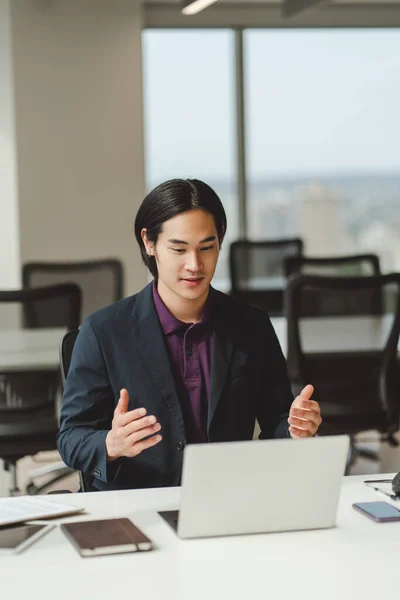 Positive asian businessman working online, having video call using laptop, sitting at workplace in modern office. Attractive japanese salesman, communication in internet. Business concept