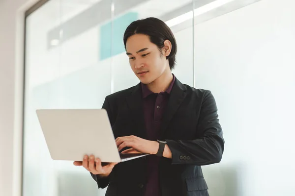 Portrait of attractive asian man standing in modern office holding, using laptop, working online. Handsome japanese manager wearing formal wear stylish suit. Concept of technology, successful business