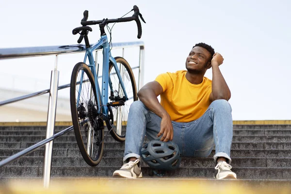 Dreamy happy african american man looking away while relaxing at the stairs after bicycle riding. People lifestyle concept