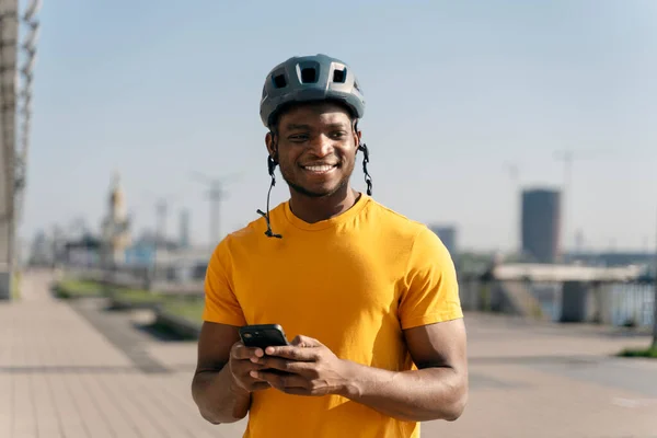 Portrait of smiling positive African man wearing safety helmet holding mobile phone on urban street. Young handsome guy wearing yellow t shirt checking mail, looking away. Technology concept