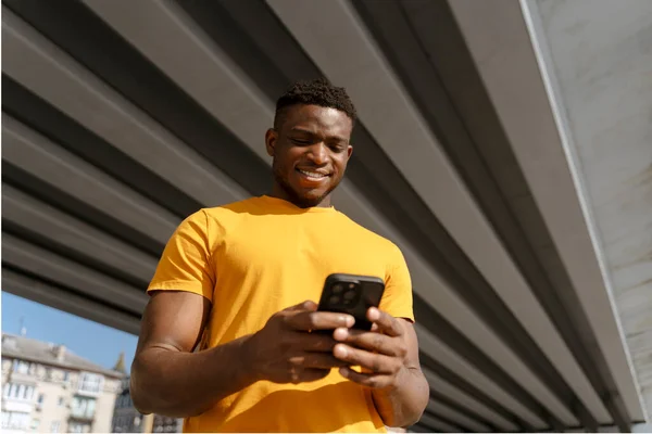 Positive young african american man holding mobile phone, watching video on city street. Handsome smiling African businessman checking mail. Concept of online communication