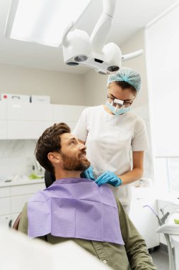 Çekici, gülümseyen bir erkek hastanın portresi dişçi koltuğunda oturmuş dişçiyle konuşuyor. Dürbünlü çekici kadın doktor diş peçetesi takıyor. Tedavi, diş bakımı kavramı