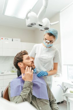 Diş sandalyesinde oturan, yanağına dokunan ve diş ağrısı çeken yakışıklı bir erkek hastanın portresi. Modern klinikte dişçiyi ziyaret eden çekici bir adam. Diş tedavisi, diş bakımı, sağlık hizmetleri