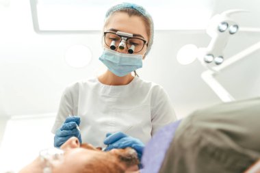 Lateks eldivenli kadın dişçi ve üniformalı modern klinikte dişçi koltuğunda yatan erkek hastanın dişlerini inceliyor. Diş sağlığı konsepti 