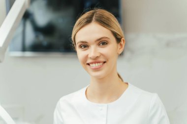 Diş muayenehanesinde durup kameraya bakan güzel, gülümseyen, sarı saçlı dişçi kadın dişçinin portresi. Modern klinikte çalışan kadın ortodontist. Diş sağlığı konsepti