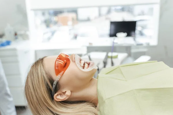 Attractive young woman with open mouth and wearing red glasses sitting in dental chair in clinic. Female patient at orthodontist consultation. Dental procedures, treatment, whitening, teeth cleaning