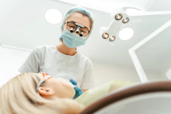 Portrait of attractive female dentist in dental binoculars examining teeth of patient sitting in chair in modern dentistry. Professional female doctor treatment, cleaning. Dental care concept