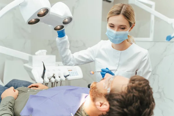 专业的女牙医在准备检查男牙的时候把灯打开 现代牙科诊所的年轻美丽微笑的女医生手持镜像设备 牙齿治疗 护理概念 — 图库照片