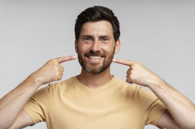 Kendine güvenen mutlu sakallı adam gri arka planda izole edilmiş dişlek gülümsemeyi işaret ediyor. Sağlık, diş tedavisi, diş hekimi. Berber dükkanından sonra kesilen şık sakallı, gülen hippi.