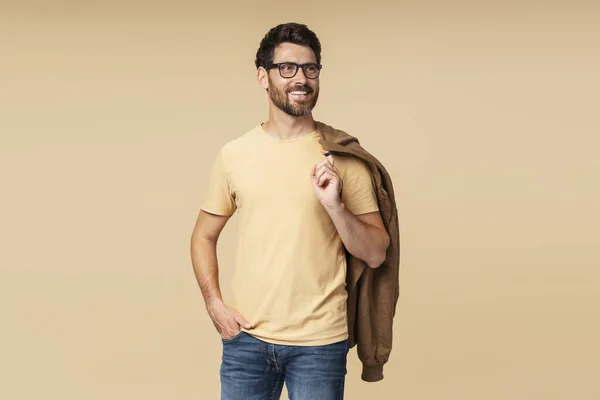 Handsome smiling bearded man holding brown autumn jacket, looking away on beige background. Portrait of successful middle aged fashion model posing for pictures, studio shot