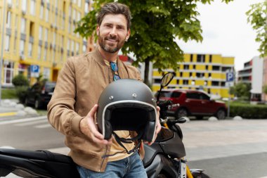 Kaskını tutan ve spor motosikletinde kameraya bakan gülümseyen bir motorcu. Yakışıklı, başarılı, şık deri ceket giyen, fotoğraf için poz veren manken.