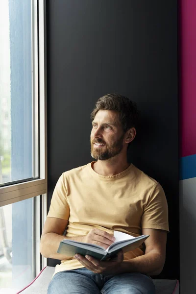 Portrait of handsome smiling man reading book sitting at home near window. Smart student studying, learning language, exam preparation in university campus. Education concept