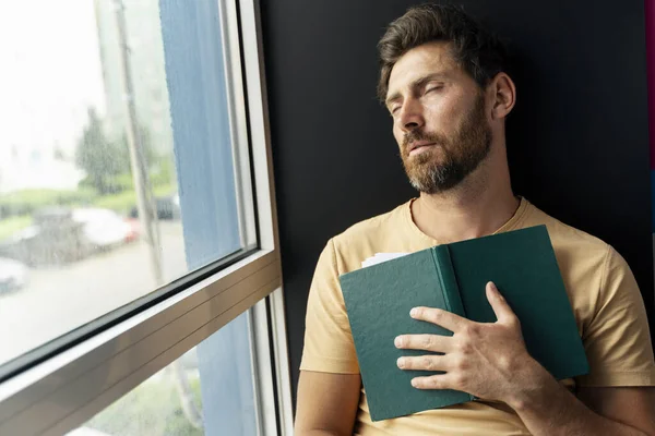 Przepracowany Śpiący Mężczyzna Brodą Trzymający Książkę Przy Oknie Portret Zmęczonego — Zdjęcie stockowe