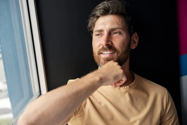 Closeup portrait of attractive happy bearded man looking at window at home. Cheerful guy rejoicing and smiling toothy alone