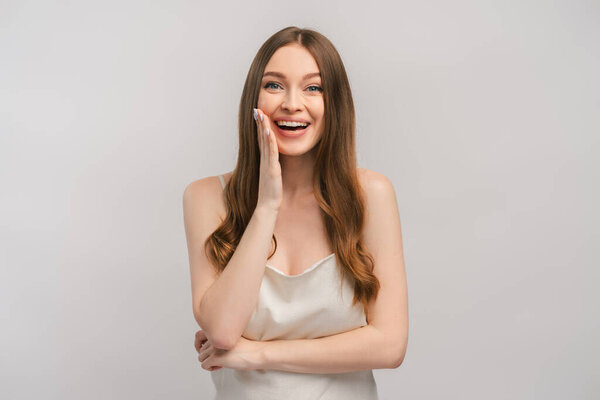 Улыбающаяся женщина в платье чувствует себя счастливой и удивлённой, держа руку на лице, глядя в камеру. Крытая студия, снятая на сером фоне