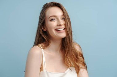Uzun, sağlıklı saçları olan, dişlek gülüşlü, mavi arka planda izole edilmiş kameraya bakan güzel bir kadının portresi. Doğal güzellik, cilt bakımı kavramı