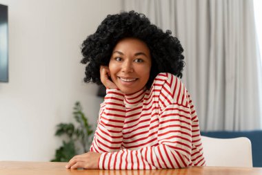 Çekici gülümseyen Afro-Amerikan kadın portresi günlük giysiler içinde masada oturmuş kameraya bakıyor. Duygusal açıdan güzel, pozitif kadın poz veriyor.