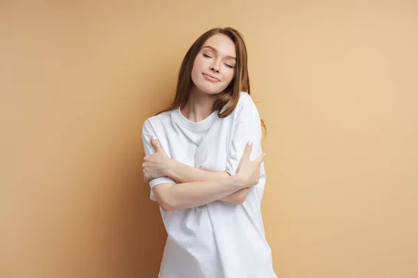 Portrait of cute woman hugging herself with closed eyes isolated on beige background. Beautiful female wearing white t shirt posing in studio