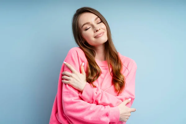 Portrait of cute woman hugging herself with closed eyes isolated on blue background. Beautiful female wearing pink sweatshirt posing in studio