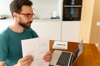 Ciddi derecede çekici sakallı, kırmızı gözlüklü, sıradan yeşil tişörtlü, iş yerinde oturan, dizüstü bilgisayar kullanan, evdeki belgelerle uğraşan bir adamın portresi. Çevrimiçi çalışan serbest çalışan, notlar alan