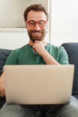 Çekici, gülümseyen sakallı beyaz adam, dizüstü bilgisayar kullanan serbest çalışan, kanepede oturan, evden internetten çalışan. Postaları kontrol eden gözlüklü yakışıklı bir programcı. Teknoloji kavramı, uzaktan çalışma