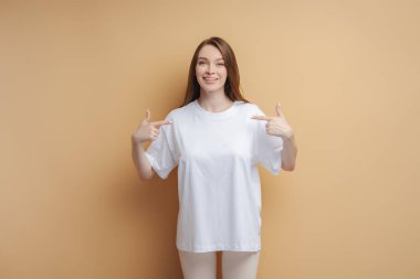 Beyaz tişörtlü güzel bir kadın bej arka planda izole edilmiş kameraya bakıyor. Model 