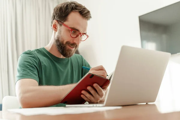 有魅力的留胡子作家的画像 他拿着笔记本和钢笔 使用笔记本电脑 坐在家里工作 做笔记 自由职业者穿着休闲绿色T恤 眼镜在网上工作 远程工作 — 图库照片