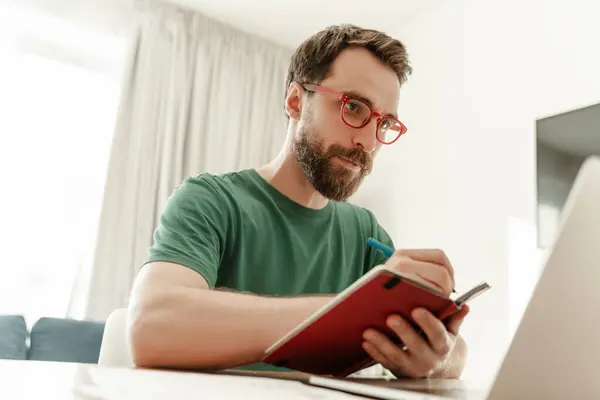 吸引认真的自由职业者 他们带着眼镜和休闲装坐在工作场所 他们在家里的客厅里拿着笔和笔记本记笔记 留胡子的男生准备考试 — 图库照片