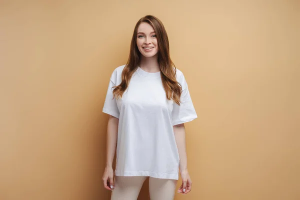 Portrait of cute smiling woman with long silky hair wearing white t shirt looking at camera isolated on beige background. Mockup