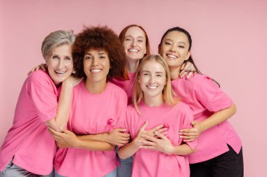 Bir grup gülümseyen kadın pembe arka planda pembe kurdeleli tişörtler giyiyor. Göğüs kanseri