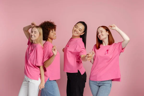 Group of smiling multiethnic women with pink ribbon dancing, celebration isolated on pink background