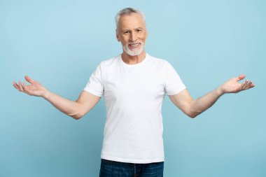 Gülümseyen emeklinin portresi kollarını açıyor ve mavi arka planda izole edilmiş kameraya bakıyor. Pozitif emeklilik aktif yaşam tarzı, reklam