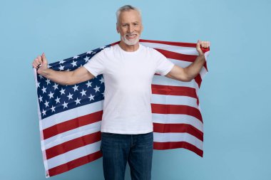 Yakışıklı, gri saçlı, yaşlı adam mavi arka planda Amerikan bayrağı tutuyor. 60 yıllık mutlu Amerikan vatanseverlik kutlaması. Kameraya bakarak Bağımsızlık Günü 'nü kutluyor. 4 Temmuz