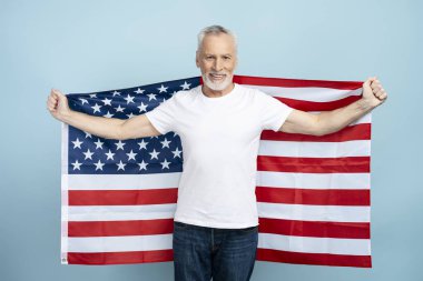 Mavi arka planda Amerikan bayrağı tutan yaşlı gri saçlı adamın portresi. 60 yaşında, beyaz tişört giyen, vatansever, model.