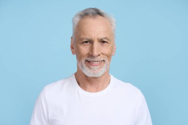 Beyaz saçlı, sakallı, yakışıklı, son sınıf öğrencisi. Mavi arka planda izole bir şekilde kameraya bakıyor. Olgun yetişkin erkek emeklinin yakın plan stüdyo portresi, olumlu duygular ifade ediyor..