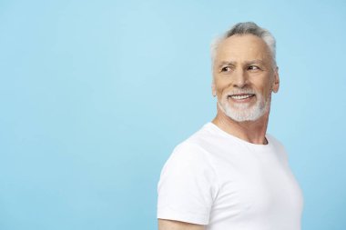 Yakından bakınca, beyaz tişört giyen, beyaz saçlı, çekici bir adam mavi arka planda tek başına, kopya boşluğuna bakıyor. Stüdyoda fotoğraf için poz veren kendinden emin bir erkek. Sağlık hizmeti kavramı