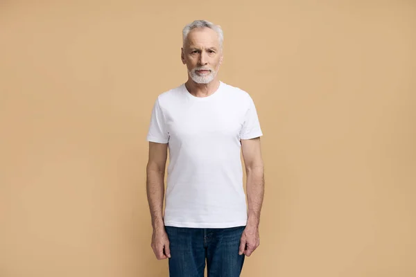 年事已高的60岁至69岁的白发苍苍的老年男子 身穿白色T恤和休闲蓝色牛仔裤 自信地看着相机 与米色背景隔离 — 图库照片