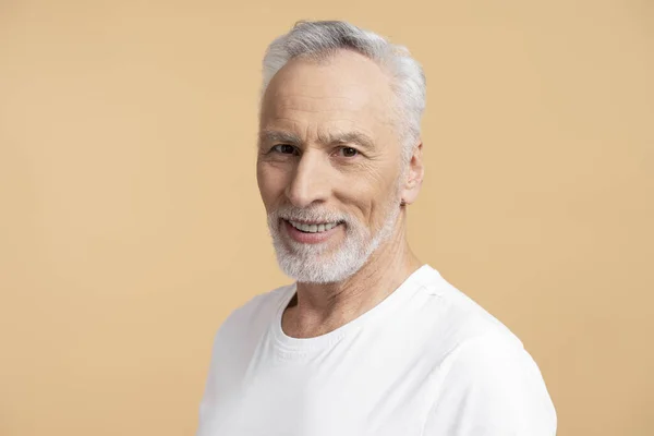 60岁的白发男子身穿白色T恤 凝视着被米色背景隔离的相机 老男人摆姿势拍照 广告概念 — 图库照片