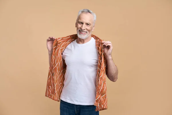 白种人灰白的头发 留着胡子的积极的老人 穿着休闲的橙色衬衫和牛仔裤 微笑着看着相机 孤立的米色背景 复制空间 积极的情绪退休人员的概念 — 图库照片