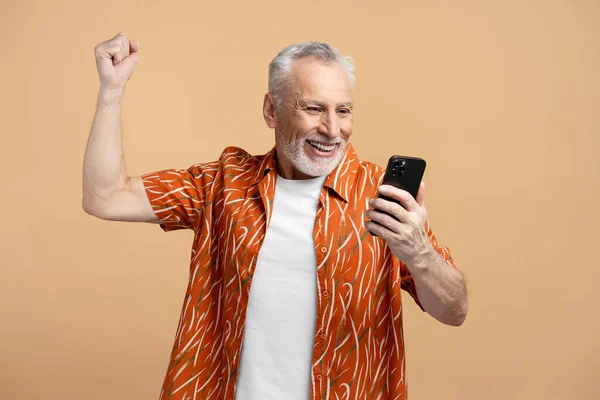 笑容可亲 头发灰白的成熟男子手持手机 观看视频 在米色背景下做出孤立无援的胜利姿态 年长男性穿着衬衫检查邮件 技术概念 — 图库照片