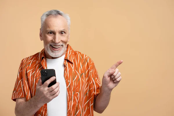 Portrait of smiling excited gray haired senior man, holding mobile phone, using smartphone pointing finger at copy space isolated on beige background. Attractive pensioner looking at camera