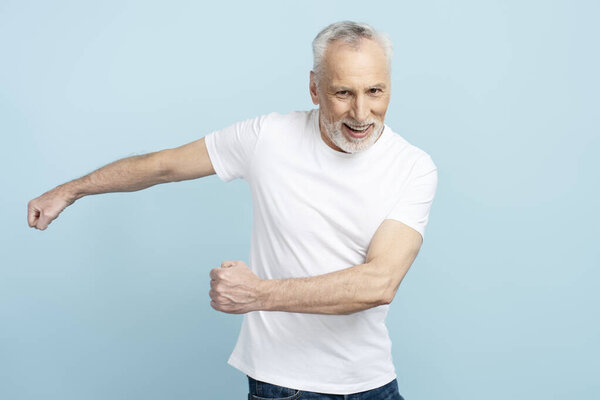 Portrait of funny smiling gray haired man showing movement gesture as if running isolated on blue background. 60 year old man wearing white t shirt, mockup